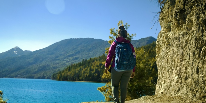Person trekking with a red bag on a dirt trail surrounded by greenery and rocky mountains.