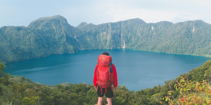Man standing on a rock with trekking gears facing the lake surrounded by greenery