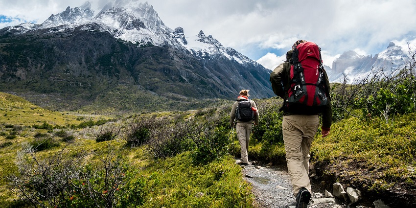 Hikers with backpacks trek a rugged mountain trail towards snow-capped peaks under clouds