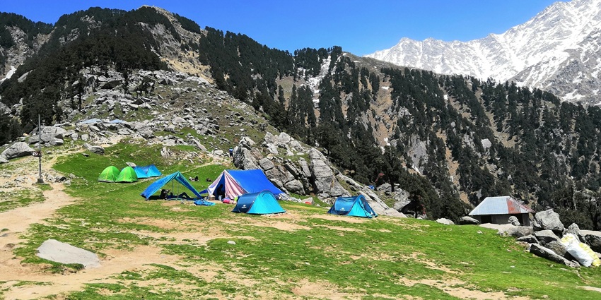 An image of people hiking on a rocky path of the Triund Trek, with snow-capped mountains and clouds in the background.