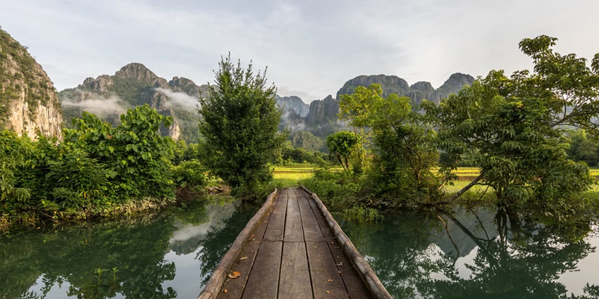 A wooden footover bridge over a lagoon with a backdrop of scenic mountains.