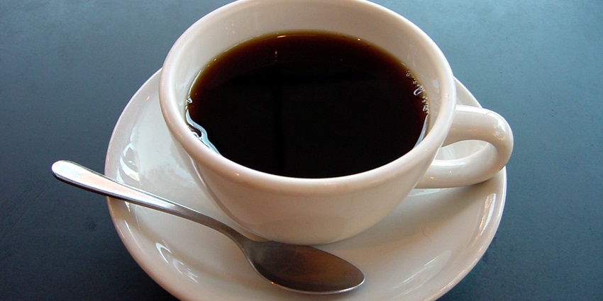 A small white cup filled with black coffee on a matching saucer, set against a neutral background.