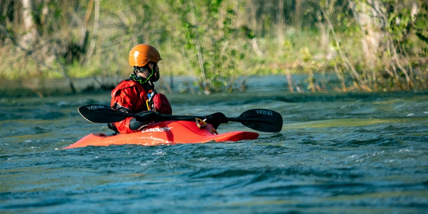A person kayaking in an orange kayak in a lake, wearing a red helmet and a safety jacket.