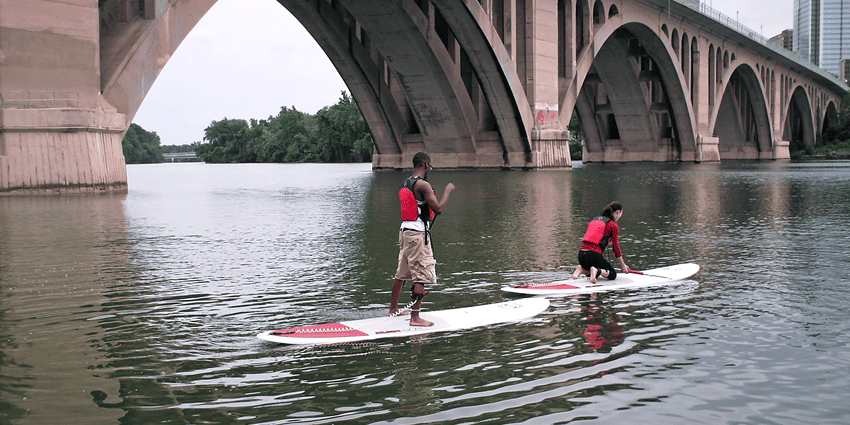 Two people stand-up paddleboarding on a wide river near a bridge under a clear sky.
