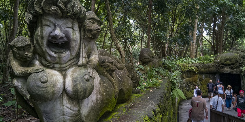 Visitors walk past mossy stone statues and monkeys in Ubud's Sacred Monkey Forest, Bali