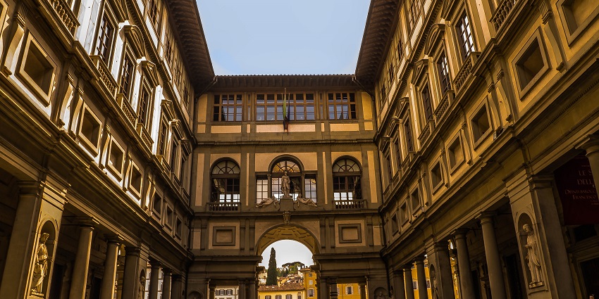 The exterior of the Uffizi Gallery in Florence shows its long courtyard framed by classical arches and statues under a clear sky.