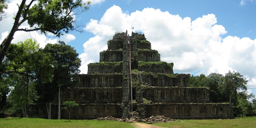Image of an ancient stepped Koh Ker Temple surrounded by trees, featuring central stairs and grassy paths.