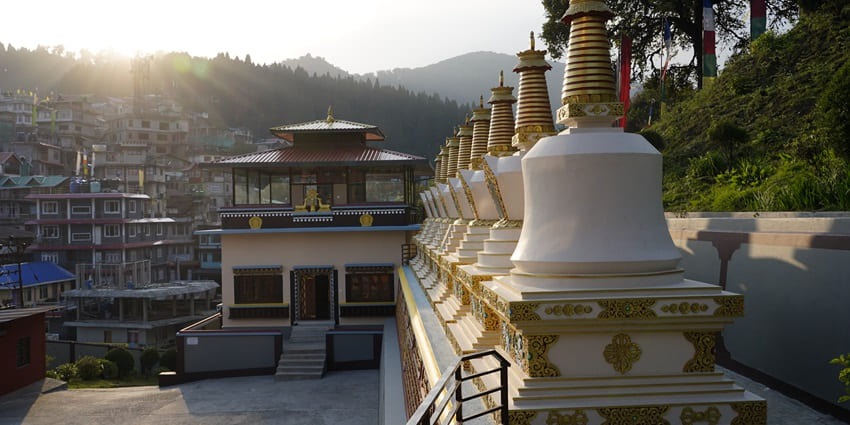 An image of a monk in red and yellow robes standing before a beautifully decorated building's staircase