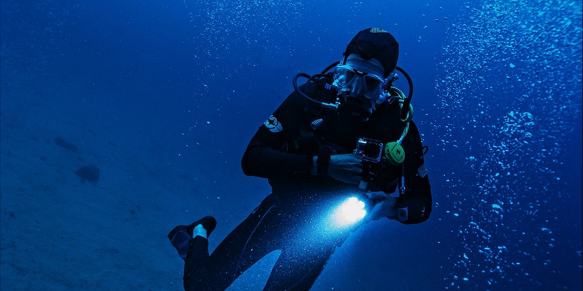 Man diving under blue waters, holding a torch, diving gear and an oxygen cylinder pipe