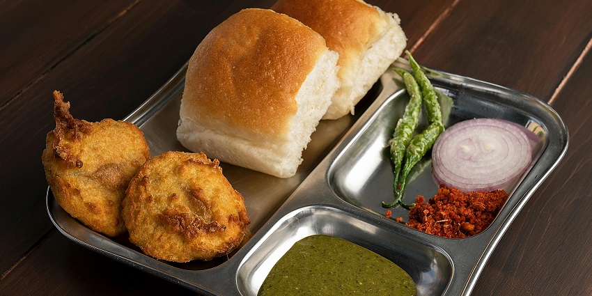An image of a plate of Vada Pav, the most famous street food in India, with a chili pepper, and a pile of chutney powder