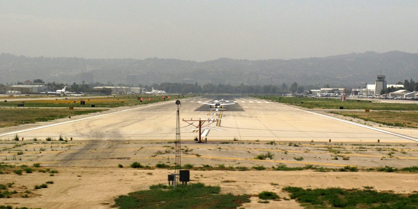 Runway of the Van Nuys Airport with distant aeroplanes parked and distant buildings.