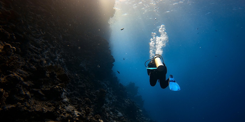 Diver with blue fins exploring a deep coral reef wall underwater, bubbles rising above.