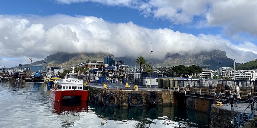 An image of the aerial view of the V&A waterfront featuring large ships, smaller boats, docks, industrial buildings, and background fog.