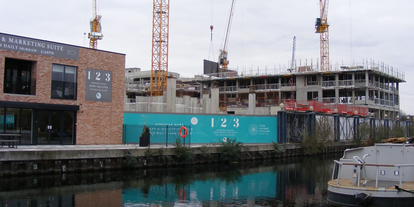 Site with cranes and partially built structures along a canal under an overcast sky.