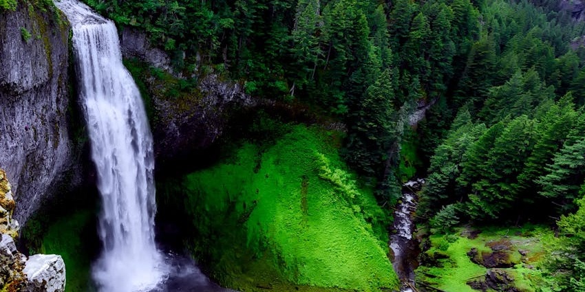 An image of the Kune waterfall cascading down a rocky cliff into a green forested area below.