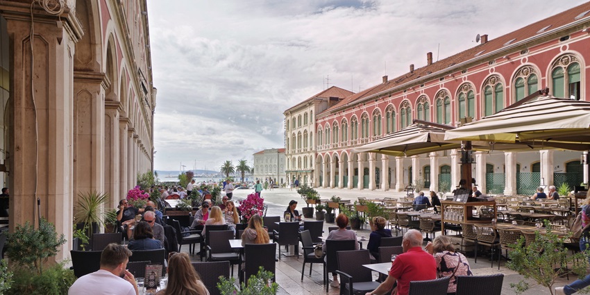 Neo-Renaissance square in Split with red façades, arched colonnades, and people relaxing at outdoor cafés under a sunny sky.