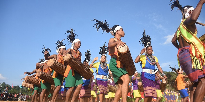 An image of individuals in colorful attire, performing the traditional dance, Wangala, with drums outdoors under clear skies.