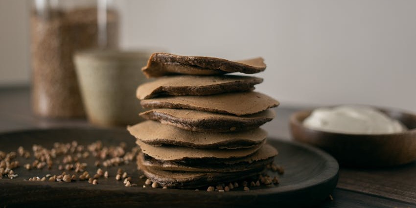 Pile of buckwheat pancakes with small seeds served on a wooden plate and other containers.