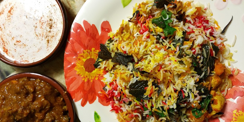 Plate of vegetable biryani with raita served in bowls on a floral plate and rustic table.