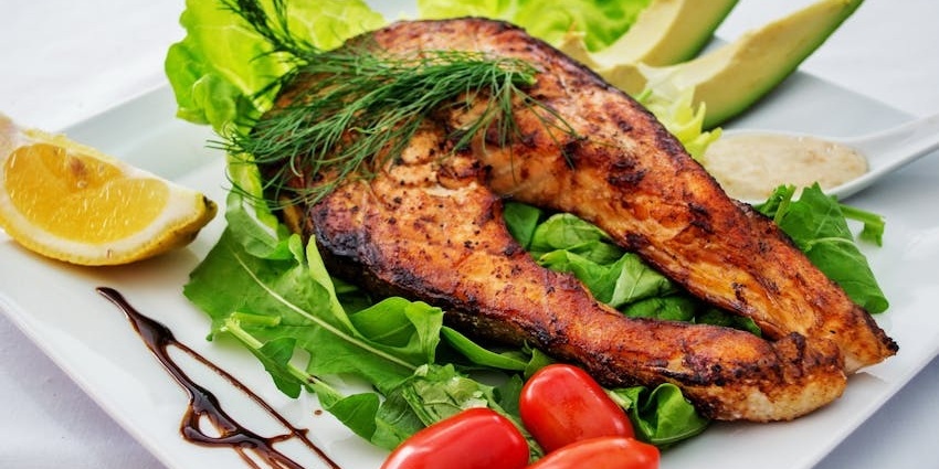 A salmon on a plate, with a lemon wedge, avocado slices, cherry tomatoes, and greens.