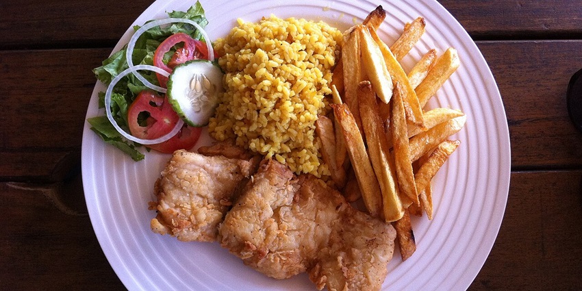 Dish in with yellow rice, potato fries, fried chicken and salad served on a white plate