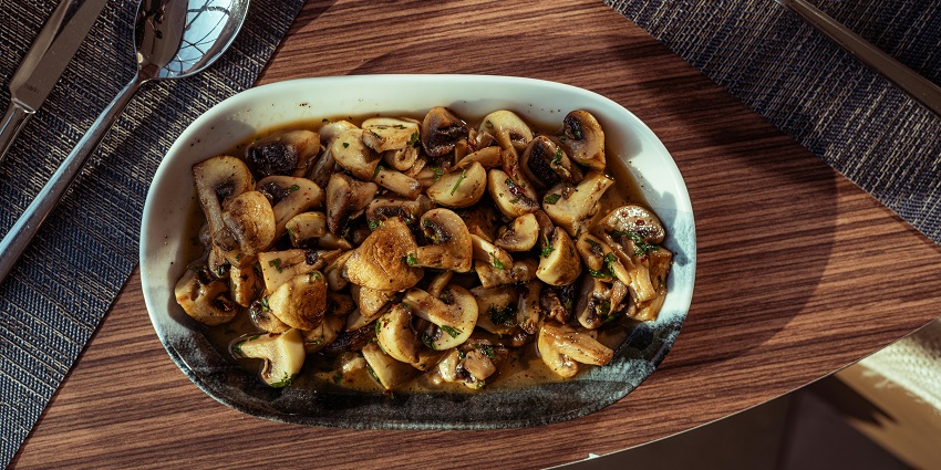 Image of a small, white, oval dish filled with sautéed mushrooms on a wooden table