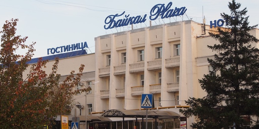 Entrance to the Baikal Plaza Hotel with blue sign boards and multiple vehicles parked.