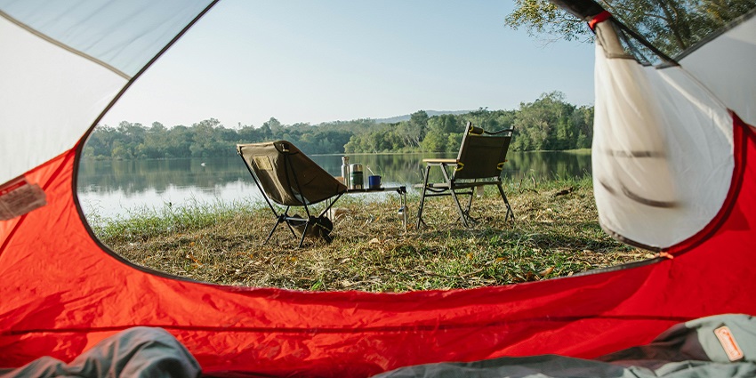 Red and white camp tent with a lake in the back, two chairs, bottles and cups on tables