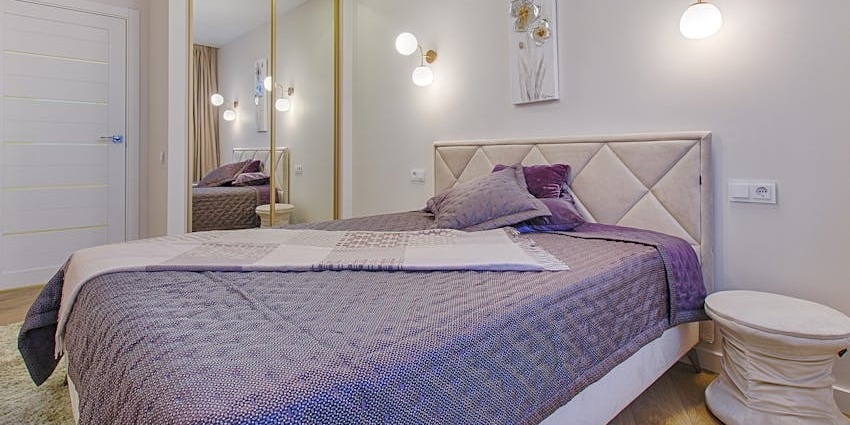 A small, modern bedroom with a neatly made bed featuring a quilted purple comforter