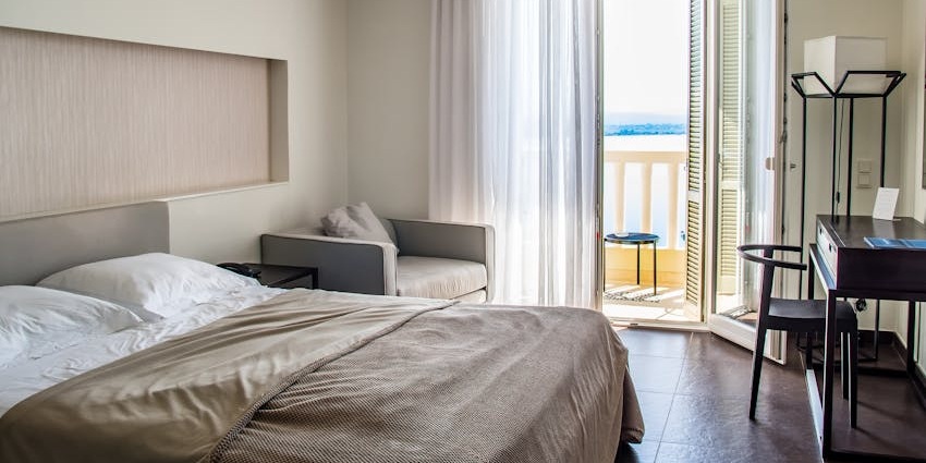 Interior of a hotel room with a bed, a lamp hanging near the bed, a window and chairs outside