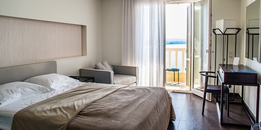 An image of a hotel room with a neatly made bed and a window leading to the balcony