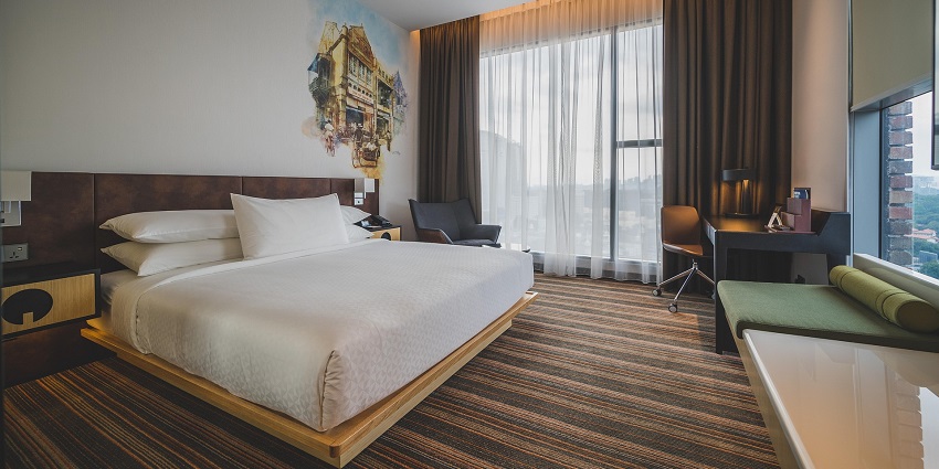 An image showing a hotel room with a bed, side tables, and a painting in the background.
