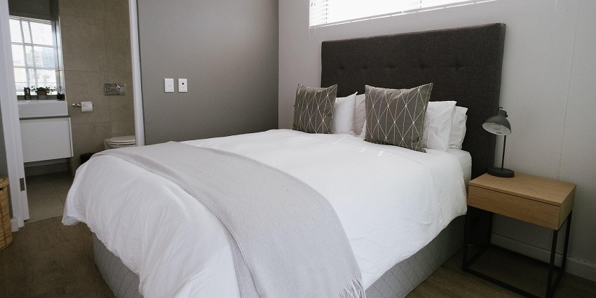An image showing a bedroom in a hotel with a neatly arranged bed and furniture around the space.