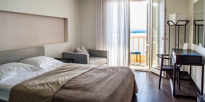 An image of a hotel room with a neatly made bed and a window leading to the balcony