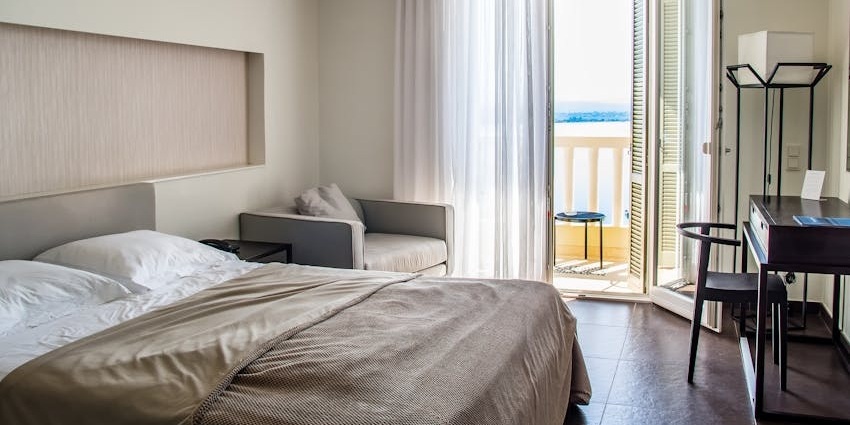An image of a hotel room with a neatly made bed and a window leading to the balcony.