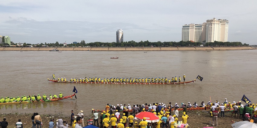 An image of a boat race on a river, during the water festival in Phnom Penh, with spectators and buildings surrounding.