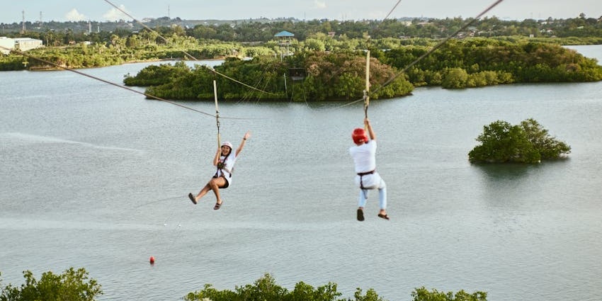 Two people waving while zip-lining over a large water body surrounded by greenery