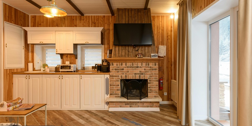 A room with a brick fireplace, a flat-screen TV, and a white kitchenette with appliances