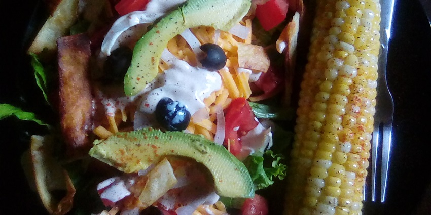 An image of a delicious Taco Salad is served with a tart corn on the cob at a restaurant.