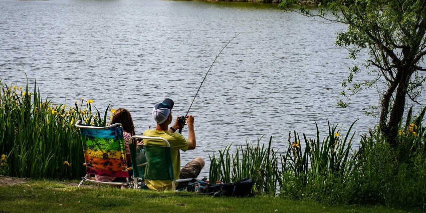 Man holding a red and black fishing rod, reeling the fishing rod and a green shoreline