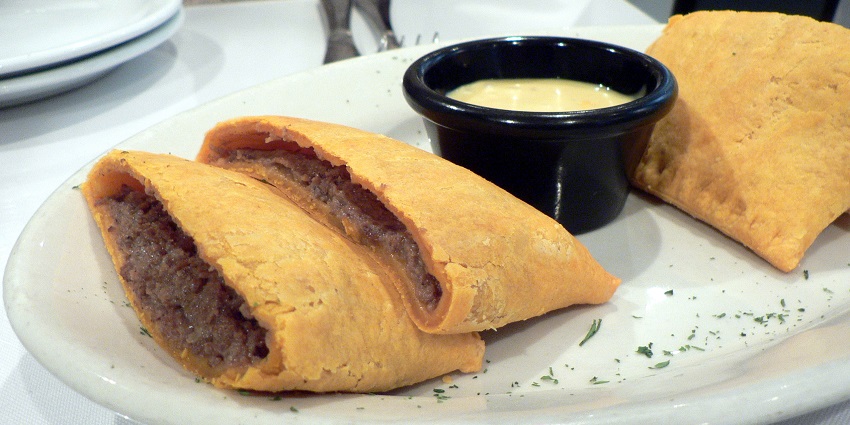 A pair of partially eaten Jamaican Beef Patties served with a bowl of sauce at a restaurant.