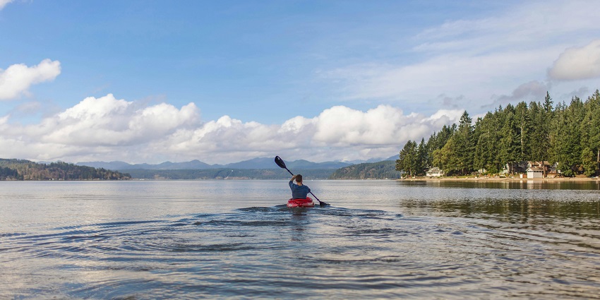 A man kayaking while holding a paddle in a large water body lake with distant mountains