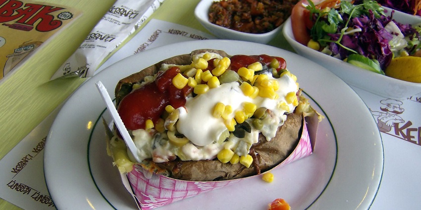 Kumpir is served loaded with cheese, sweet corn, and tomato sauce as a white spoon sticks out.