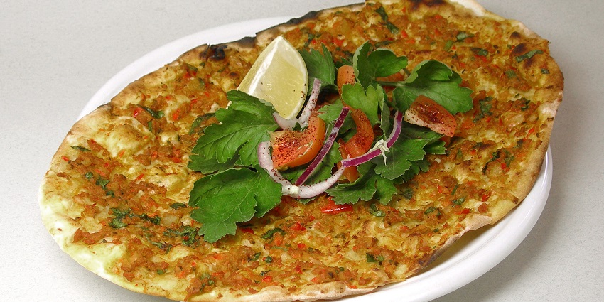 Lahmacun is served with a lime slice, green herbs, and tomato slices drizzled with chilli powder.
