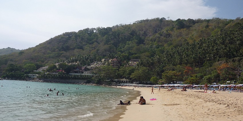 A panoramic view of Nai Harn Beach in Phuket with turquoise waters, white sand, and lush hills, showcasing a serene coastal escape in Thailand.
