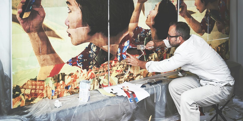 Man in white long-sleeved shirt seated indoors, focused on creating a large artistic collage.