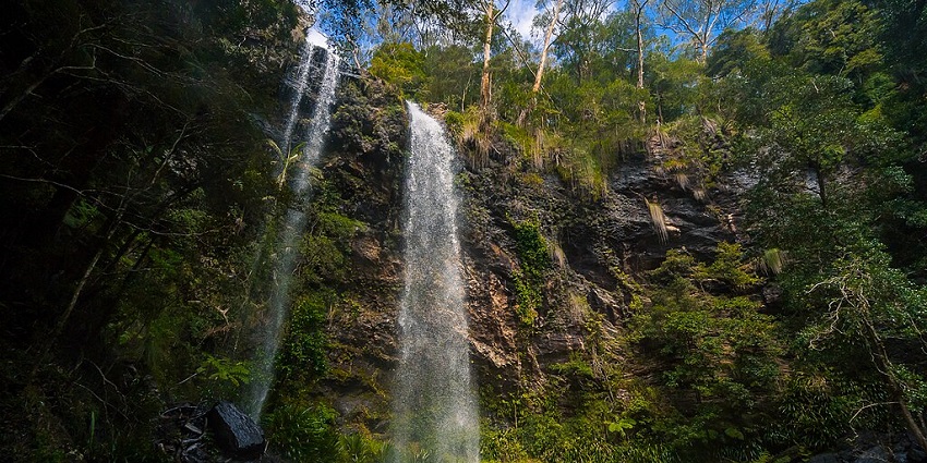 Twin waterfalls cascade down a lush green cliff into a pool, creating a picturesque scene.