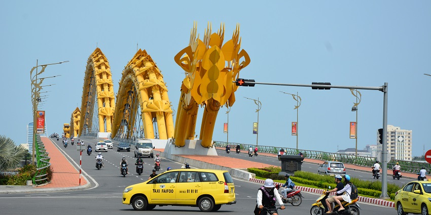 Motorbikes and cars crossing Dragon Bridge beneath a cloudy afternoon sky.