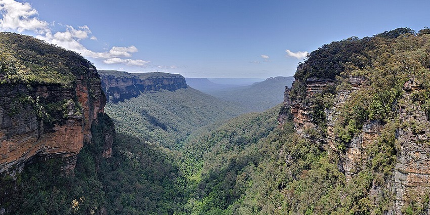Aerial view of a lush green valley with steep cliffs and dense forest under a clear sky.