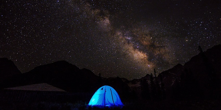 A dreamy view of people camping in a blue tent under a blanket of glittering stars at night